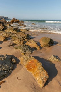 A rocky beach with a large rock in the foreground and a boat in the distance. The beach is quiet