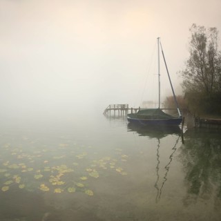 Quiet autumn morning on Lake Chiemsee, jetties and sailing boat in dense fog, water lilies in the