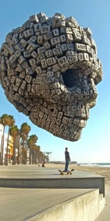 A person on a skateboard is captured at a seaside boardwalk, with the shore and a striking skull