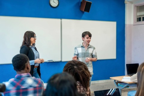 Teenage student giving a presentation in a modern classroom with a teacher and diverse group of