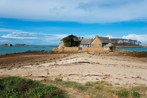 An isolated stone house stands on a small promontory next to the sea under a blue sky, house at