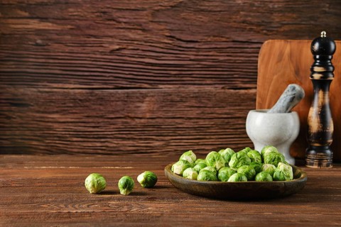 A wooden bowl filled with fresh Brussels sprouts sits on a rustic wooden table. Nearby, a mortar,