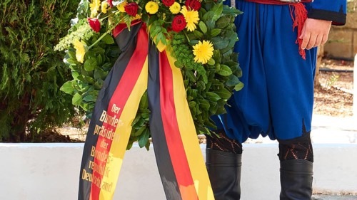 Wreath of flowers in the colours of Germany with a man dressed in traditional Cretan costume, visit