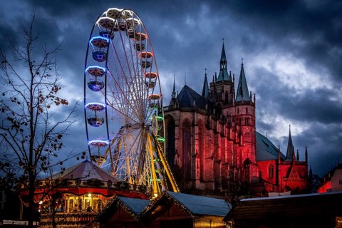 Ein beleuchtetes Riesenrad vor dem Hintergrund einer Kathedrale und einem dunklen Nachthimmel,