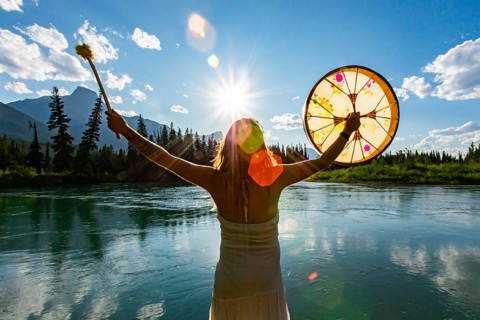 A close up and rear view of a shaman woman holding a sacred powwow drum towards sun and mountains