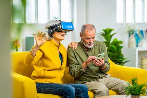 Senior woman using virtual reality glasses next to her husband sitting on the sofa using mobile app