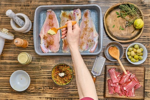 Fresh fish fillets are being seasoned in a rustic kitchen. Herbs, spices, and olive oil are neatly