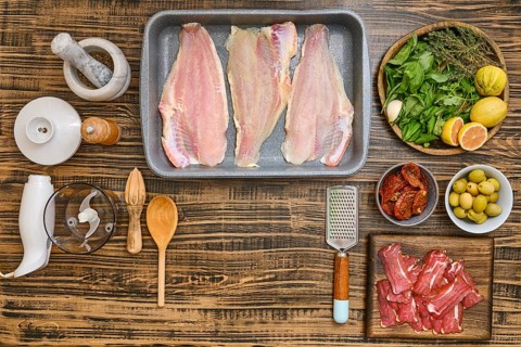 Various fresh ingredients are arranged on a wooden tabletop, including fish fillets, herbs, olives,