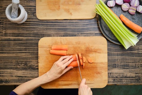 A person is chopping carrots on a wooden cutting board while surrounded by fresh vegetables,