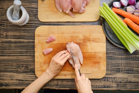 Hands skillfully cut raw chicken fillets on a wooden cutting board surrounded by fresh vegetables