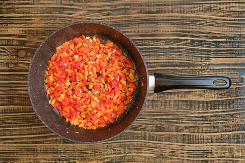 Brightly colored bell peppers are chopped and sauteing in a frying pan on a rustic wooden surface.