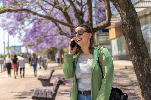 A cheerful young woman with sunglasses and a green jacket talks on her phone during a sunny day on