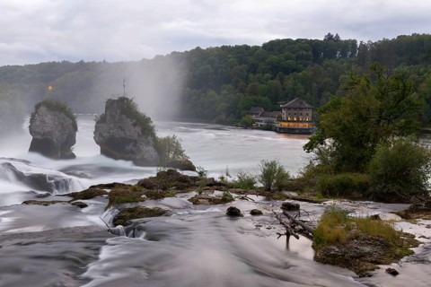 Impressive waterfall with two rocks, surrounded by forest and a building in the background in