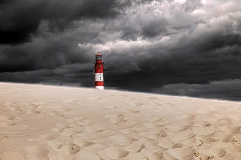 A lonely lighthouse stands on a sand dune under a threatening, grey, cloudy sky