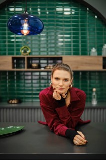 A woman is leaning on a kitchen counter, wearing a red outfit with long sleeves. The background