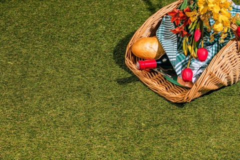 A picnic basket with bread, wine and fresh flowers lies on grassy ground