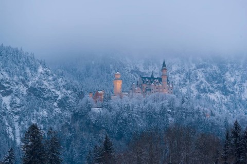 Castle rises above a quiet, snow-covered winter landscape under a misty sky, illuminated,