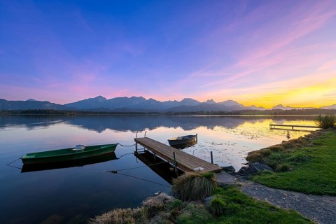 Boats at a jetty on a calm lake at sunset with mountains in the background, Hopfensee, Hopfen am
