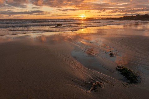 A beach with a rock and a sunset, the sky is reflected on the sand of a beach. The sky is cloudy