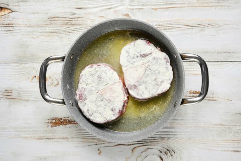 Overhead view of two floured steaks are sizzling in a pan with oil. The pan is on a white wooden