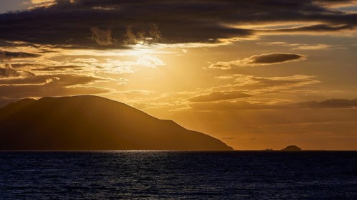 The sky shows a dramatic sunset with dark clouds and an island on the horizon, sunset, Kassos,