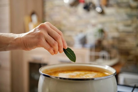 A hand reaches to place a fresh bay leaf into a pot of flavorful soup. The warm kitchen setting