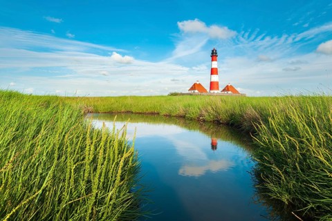The Westerheversand lighthouse under a blue sky with white clouds reflected in a tideway,