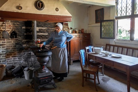 Old woman cooking on antique Flemish cast-iron coal stove, Leuvense stoof in 19th century house,