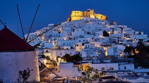 A windmill and illuminated streets lead to an illuminated fortress above white buildings, Querini