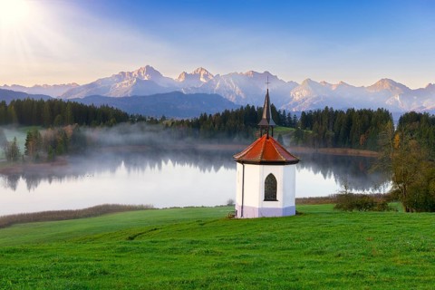 Small chapel on the lake in front of an impressive mountain backdrop at sunrise, surrounded by