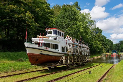A boat being transported overland on a railway wagon, surrounded by forest and blue sky, ship