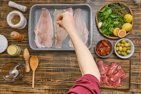 Sliced fish is placed in a baking tray while an outstretched hand sprinkles seasonings over it.