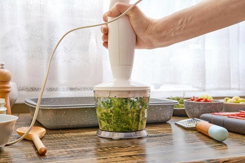 A hand holds an immersion blender blending fresh green vegetables in a warm kitchen filled with