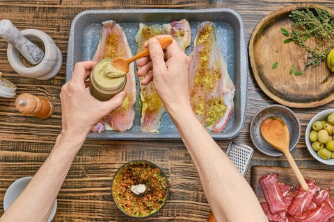 A person is mixing a spice blend in a jar while seasoning fish fillets laid out on a baking tray