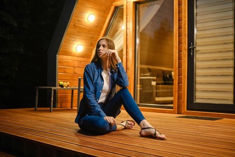 A young woman sits relaxed in the evening on a wooden terrace at home and looks thoughtfully to the