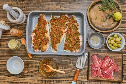 Three fish fillets are seasoned with herbs and spices, surrounded by a variety of ingredients on a
