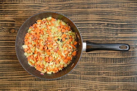 Colorful chopped carrots, onions, and celery are cooking in a black skillet on a wooden table. The