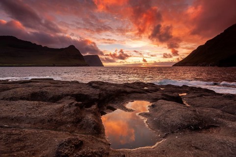 Sunset over the sea between steep mountain slopes, reflection in calm pool on rocks, Vidareidi,
