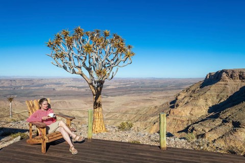 A woman sits with a cup of tea in a chair on the terrace of the Fish River Lodge, behind her the