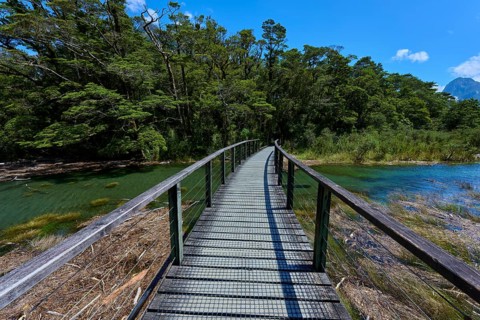 Curved wooden walkway leads over a clear river through a dense forest landscape under a sunny sky,