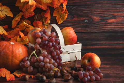 A wooden basket with apples and grapes next to chestnuts, a pumpkin and autumn leaves