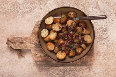 A bowl of golden roasted baby potatoes is garnished with herbs and garlic. It rests on a wooden