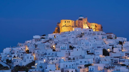 An illuminated fortress towers above white-washed buildings under a deep blue night sky, Querini