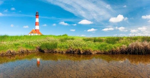 The Westerheversand lighthouse under a blue sky with white clouds reflected in a tideway,