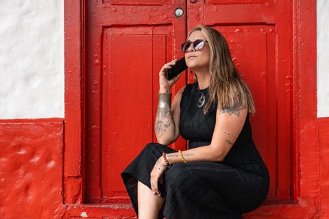 A stylish tattooed woman, dressed in a black dress and sunglasses, holds her smartphone while