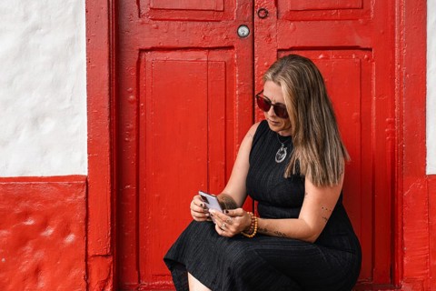 A tattooed woman in sunglasses and black dress is sitting in front of a vibrant red door,