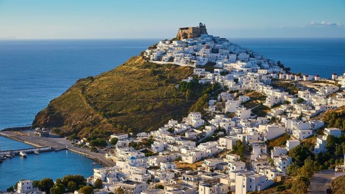 A fortress overlooks a coastal landscape of white houses gently sloping down to a blue sea, Querini