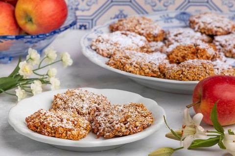 Sugar-dusted oat biscuits on a plate surrounded by apples and flowers in front of patterned tiles