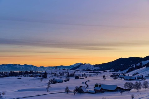 Snowy landscape at sunrise with mountains and farms, bathed in a warm orange-blue light, Menzingen,