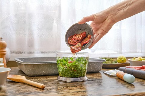 A hand pours dried tomatoes into a bowl of blender filled with vibrant green vegetables. The